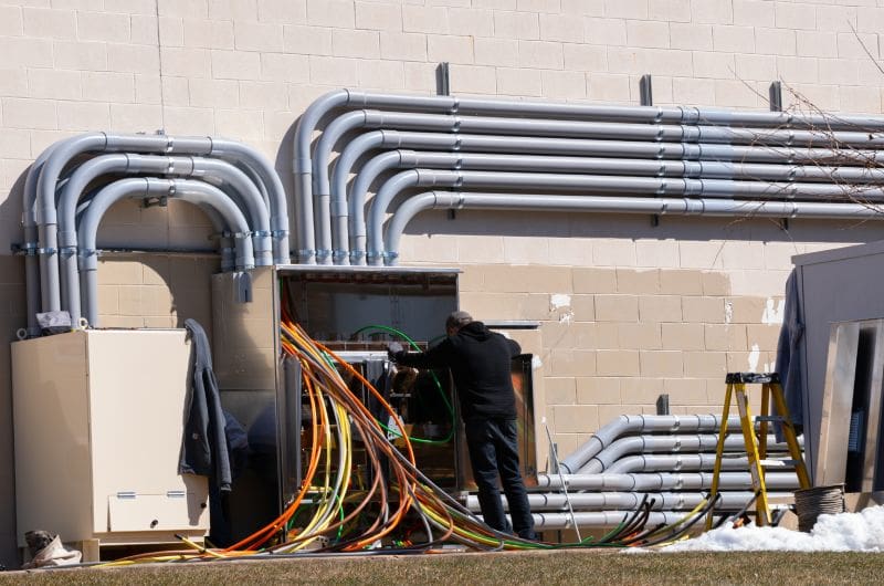 A technician works on wiring inside an open industrial electrical cabinet with multiple large conduit pipes on the exterior wall of a building.