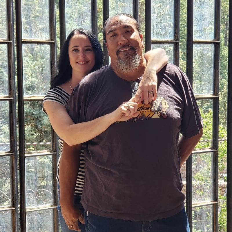 A man and a woman posing together, holding hands in front of a barred window with trees in the background.