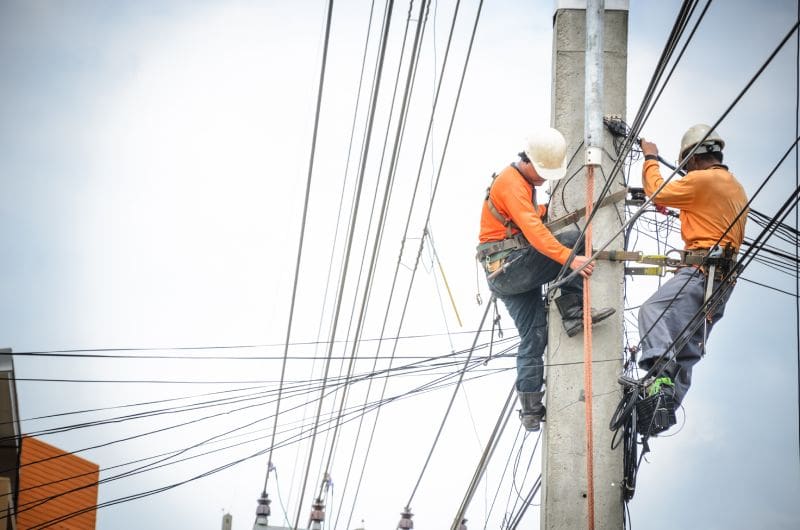 Two workers in orange safety vests and helmets install cables on a utility pole, surrounded by multiple power lines against a cloudy sky.