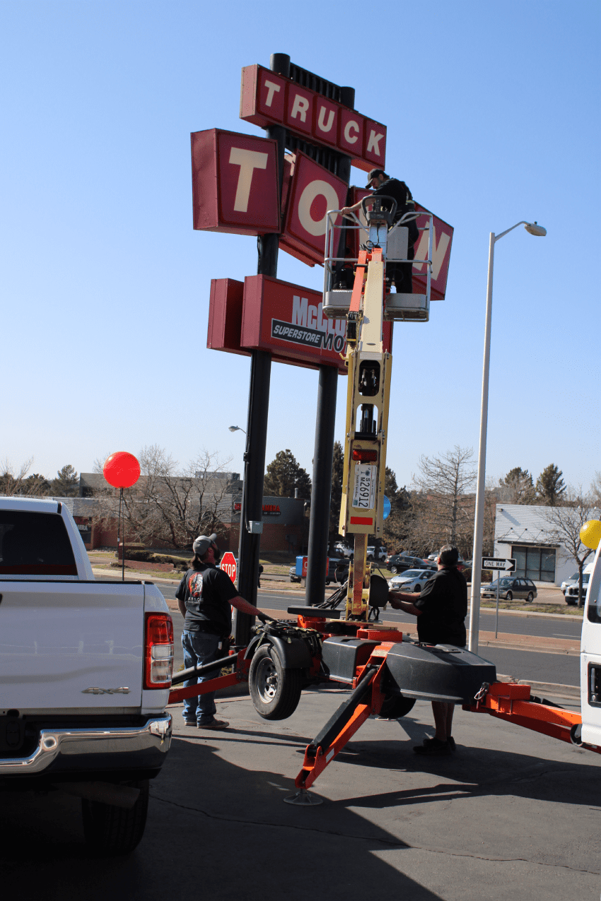 Workers use a boom lift to repair or install letters on a large roadside sign in a parking lot on a sunny day.