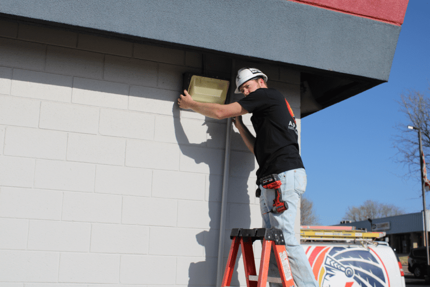 A worker in a hard hat stands on a ladder and installs or repairs an exterior light fixture on the side of a building.