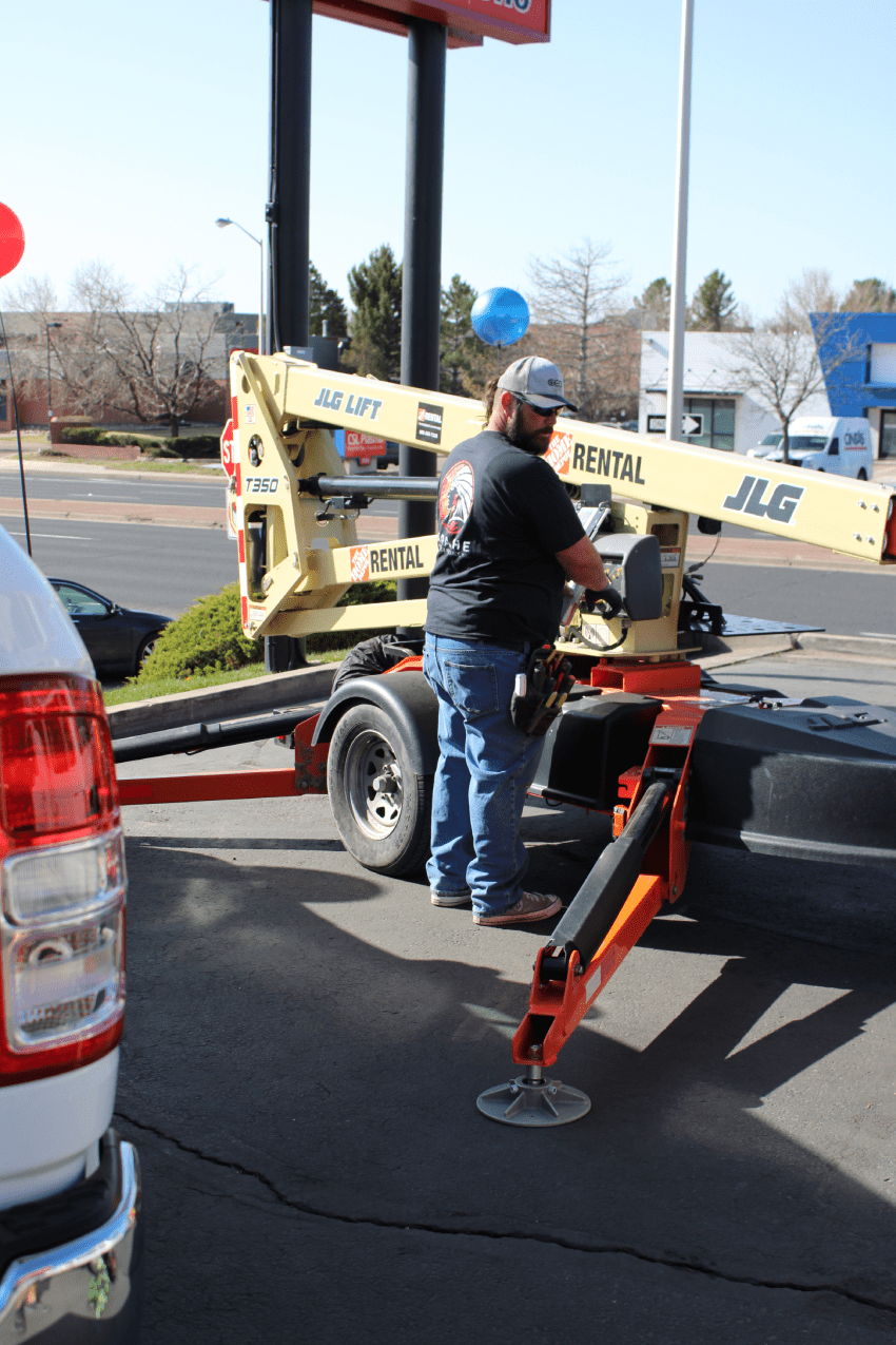 A man operates a JLG lift rental mounted on a trailer, positioned on a paved lot with vehicles and balloons visible in the background.