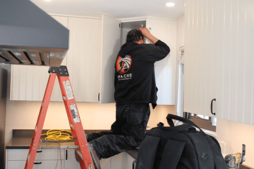 Residential custom A person on a ladder installs or adjusts a white upper kitchen cabinet; tools and a black bag are visible on the counter nearby.