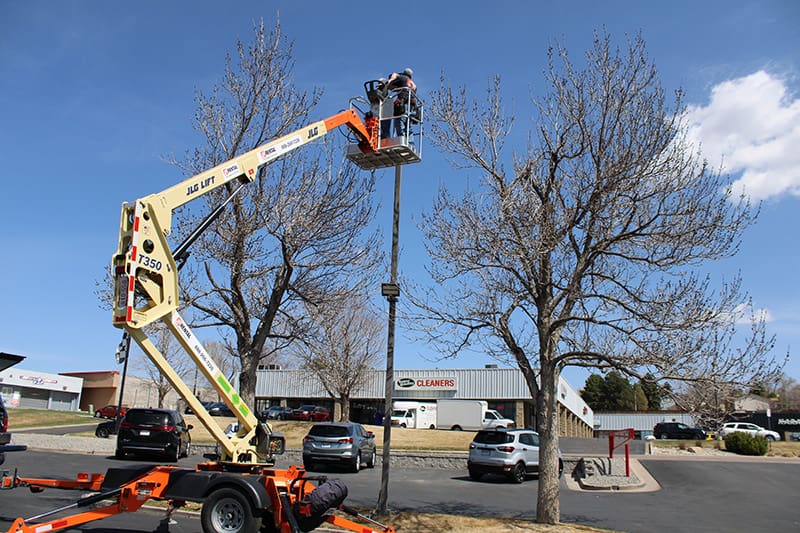 A person in a lift bucket works on a pole beside a parking lot with several cars, trees without leaves, and commercial buildings in the background.