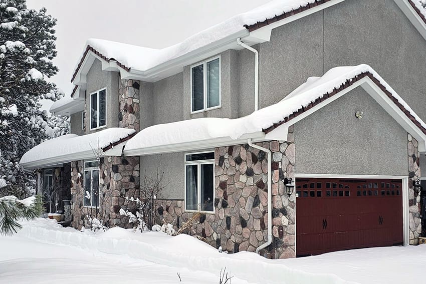 A two-story house with stone and stucco exterior is covered in a thick layer of snow on the roof, yard, and driveway.