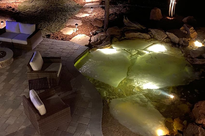 Outdoor patio with wicker chairs and a stone fire pit next to a pond illuminated by underwater lights at night. Stone pavers and landscape lighting visible in the background.