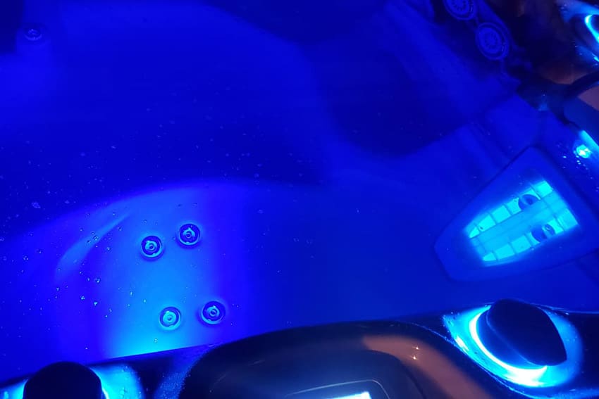 Close-up view of a hot tub interior illuminated with blue LED lights, showing control knobs, water jets, and bubbles on the water surface.