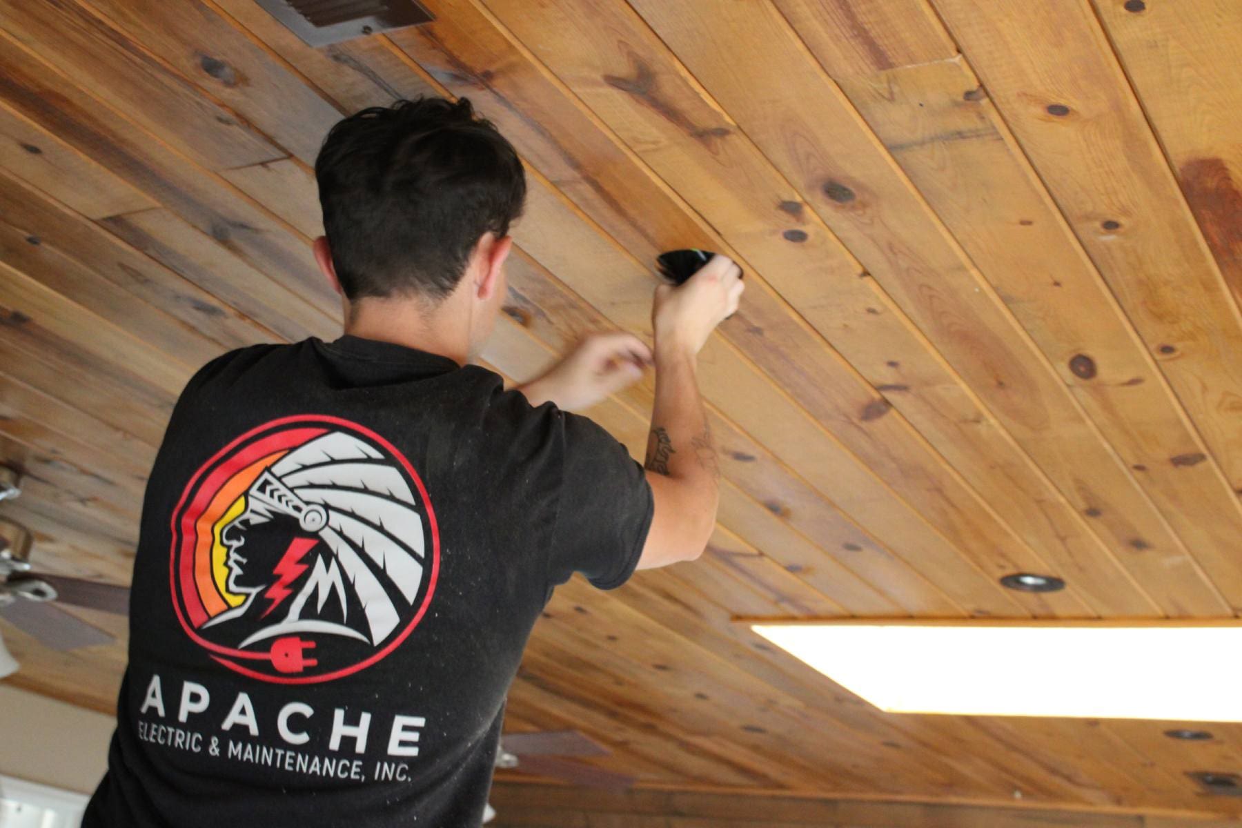 A person in an "Apache Electric & Maintenance, Inc." shirt installs or adjusts a fixture on a wooden ceiling.