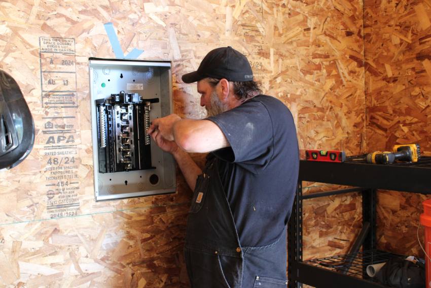 A man wearing a black cap and overalls installs or repairs wiring inside an electrical panel mounted on a plywood wall.